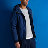 A man wearing a navy blue hooded jacket, a light gray ribbed sweater, dark navy pants, and a muted green baseball cap, standing against a solid blue studio background.