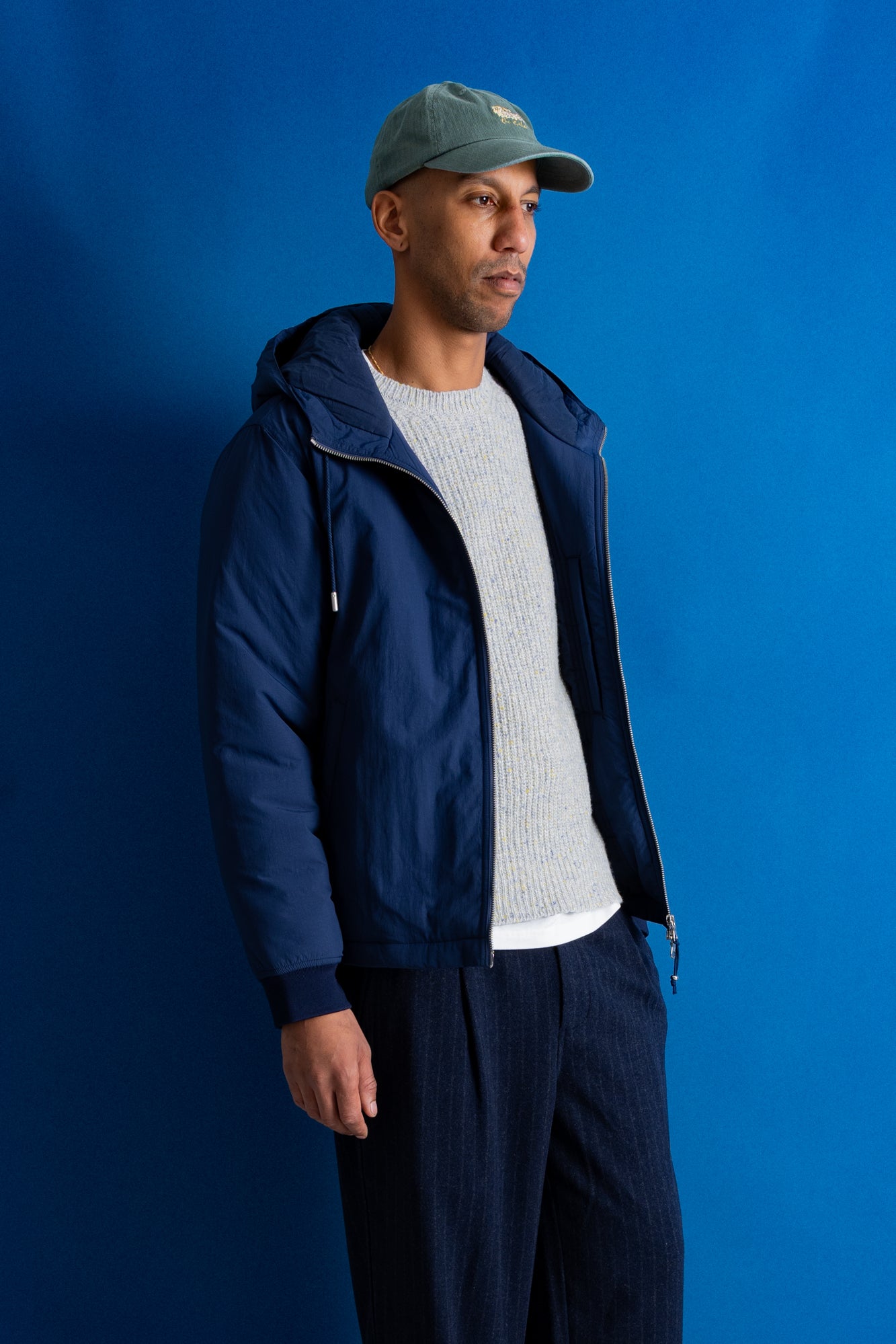 A man wearing a navy blue hooded jacket, a light gray ribbed sweater, dark navy pants, and a muted green baseball cap, standing against a solid blue studio background.
