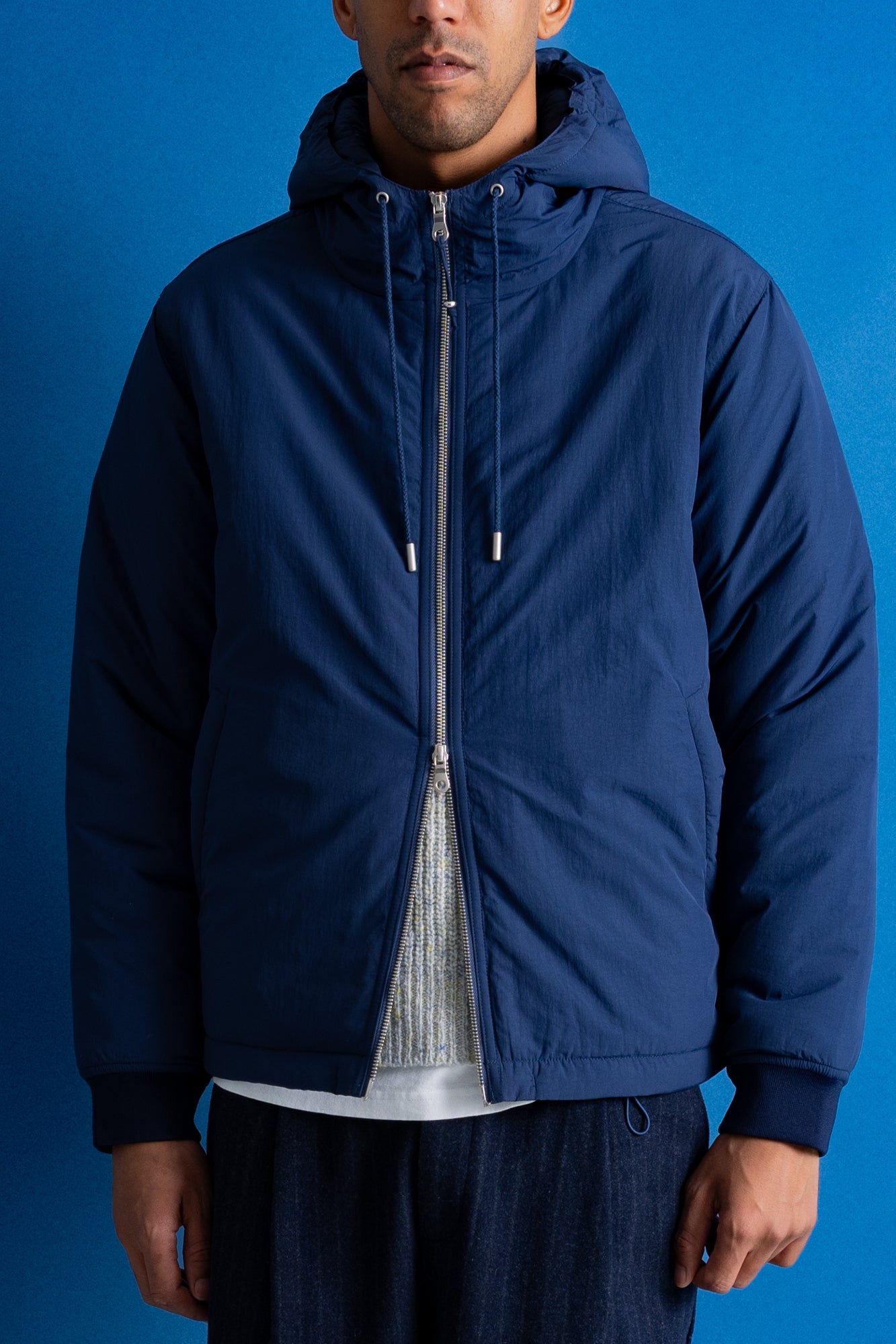 A man wearing a navy blue hooded zip-up jacket, shown from the chin down against a solid blue background.