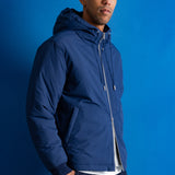 A man wearing a navy blue hooded jacket, a gray baseball cap, and dark pants, standing against a solid blue studio background.