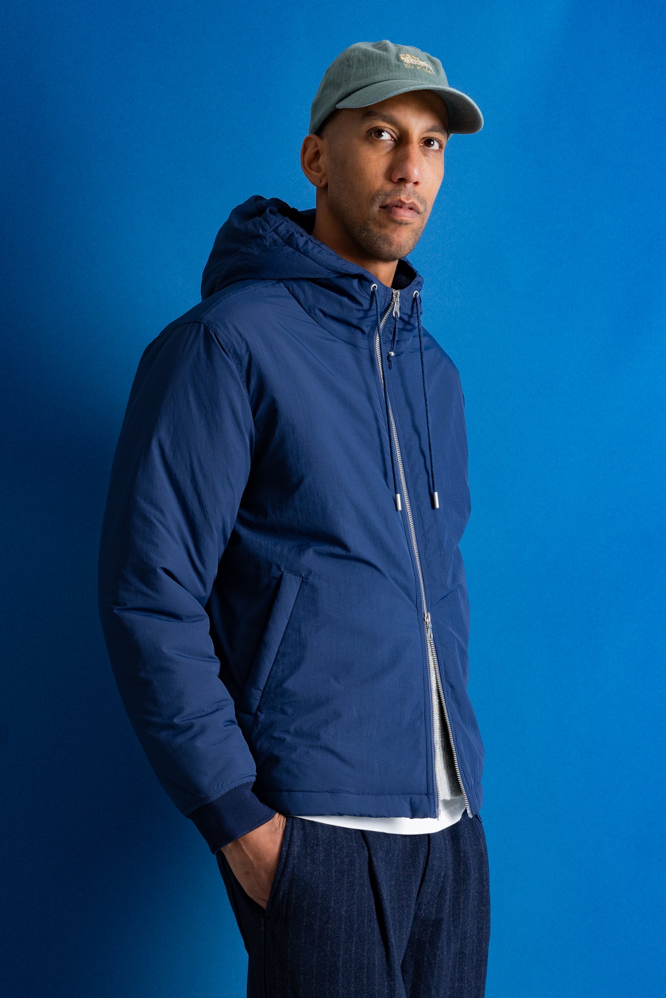 A man wearing a navy blue hooded jacket, a gray baseball cap, and dark pants, standing against a solid blue studio background.