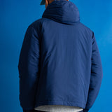 A man viewed from behind wearing a navy blue hooded puffer jacket, a gray-green baseball cap, and dark pants, standing against a solid blue studio background.