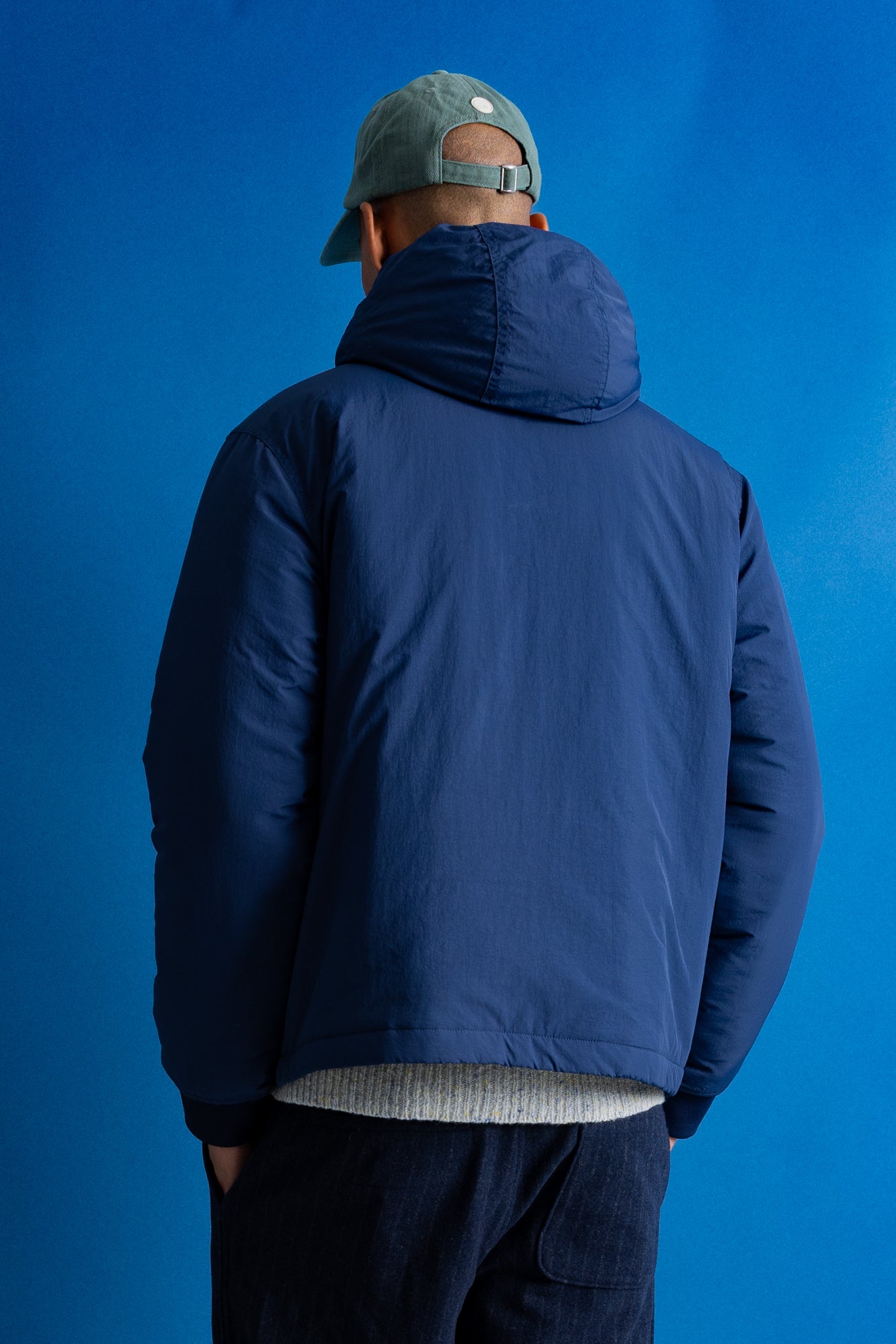 A man viewed from behind wearing a navy blue hooded puffer jacket, a gray-green baseball cap, and dark pants, standing against a solid blue studio background.