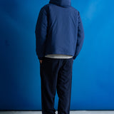 A man viewed from behind wearing a navy blue hooded jacket, dark pants, and a green cap, standing against a bright blue studio backdrop.