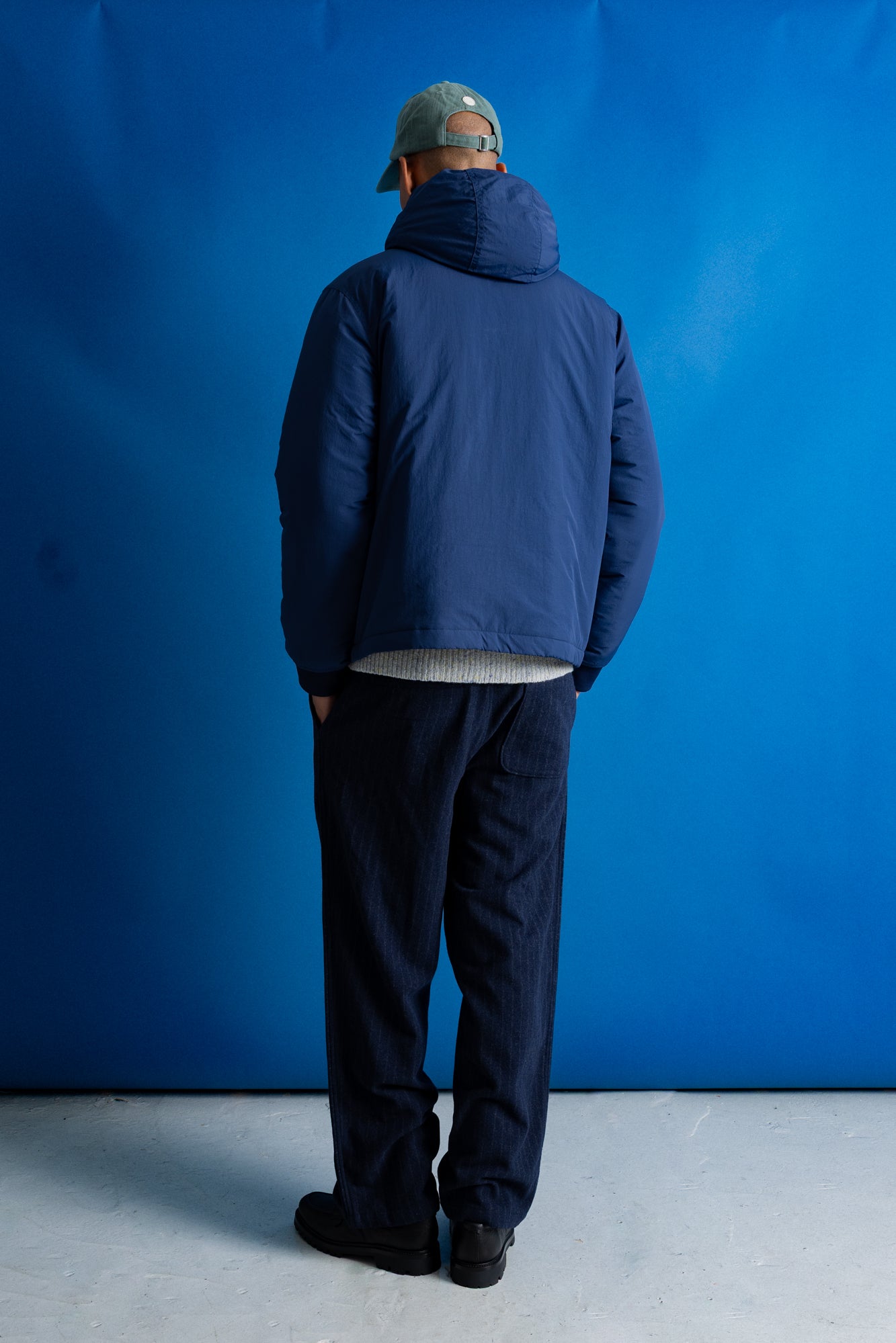 A man viewed from behind wearing a navy blue hooded jacket, dark pants, and a green cap, standing against a bright blue studio backdrop.