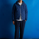 A man stands against a bright blue studio backdrop wearing a navy blue zip-up hooded jacket over a white ribbed sweater, dark navy pants, black shoes, and a light blue-green baseball cap.