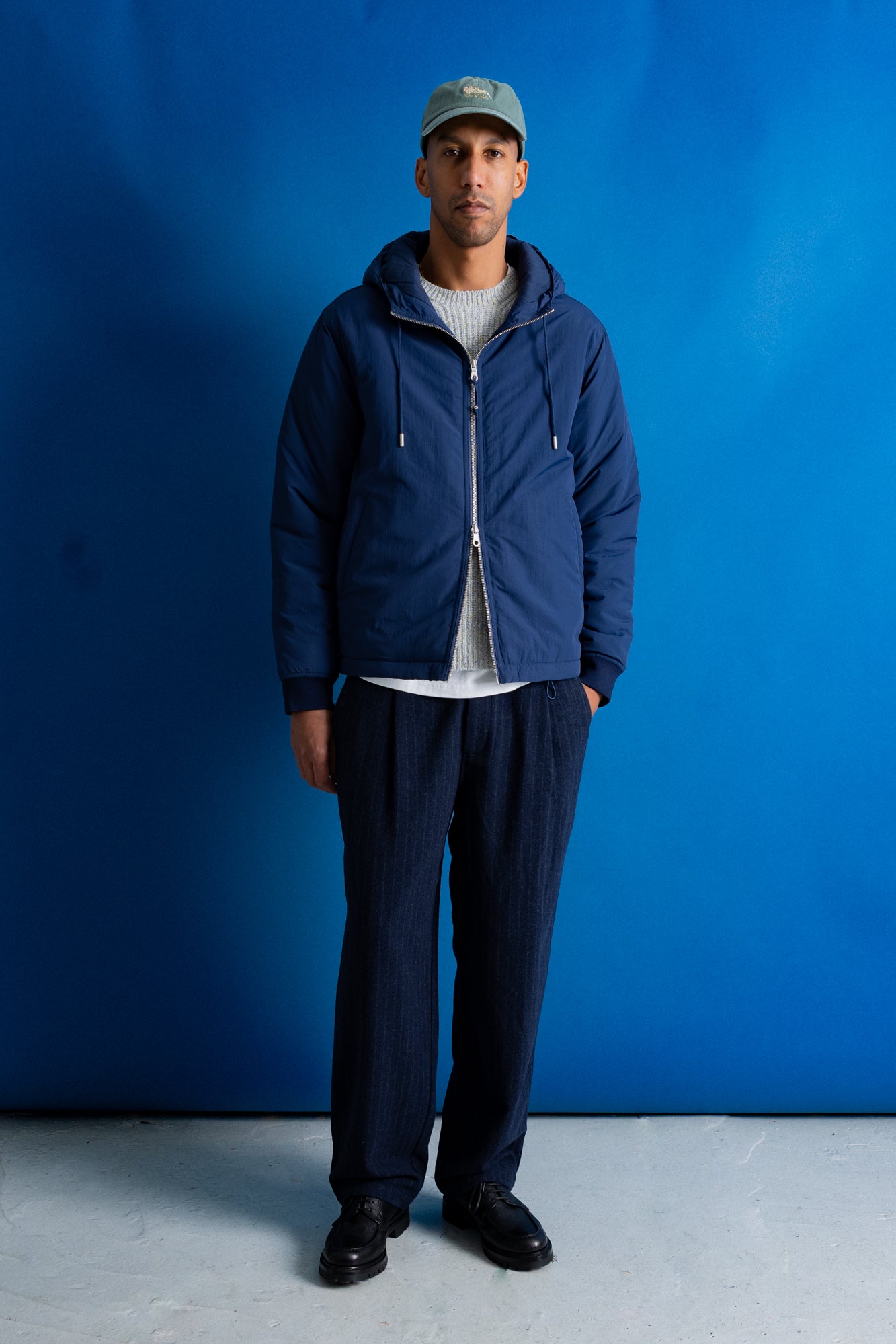 A man stands against a bright blue studio backdrop wearing a navy blue zip-up hooded jacket over a white ribbed sweater, dark navy pants, black shoes, and a light blue-green baseball cap.