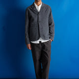 A man stands against a bright blue studio backdrop wearing a gray textured blazer over a white collared shirt, dark trousers, and black shoes.