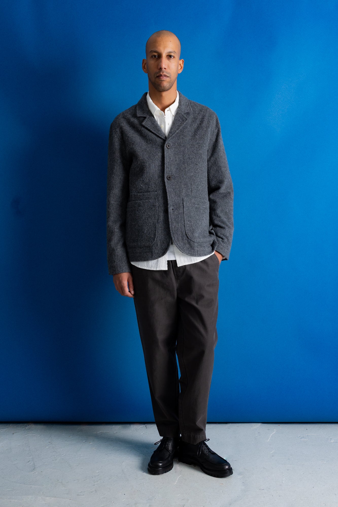 A man stands against a bright blue studio backdrop wearing a gray textured blazer over a white collared shirt, dark trousers, and black shoes.