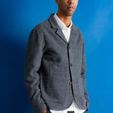 A man wearing a textured gray wool-blend blazer over a white collared shirt and dark trousers, standing against a solid blue studio background.