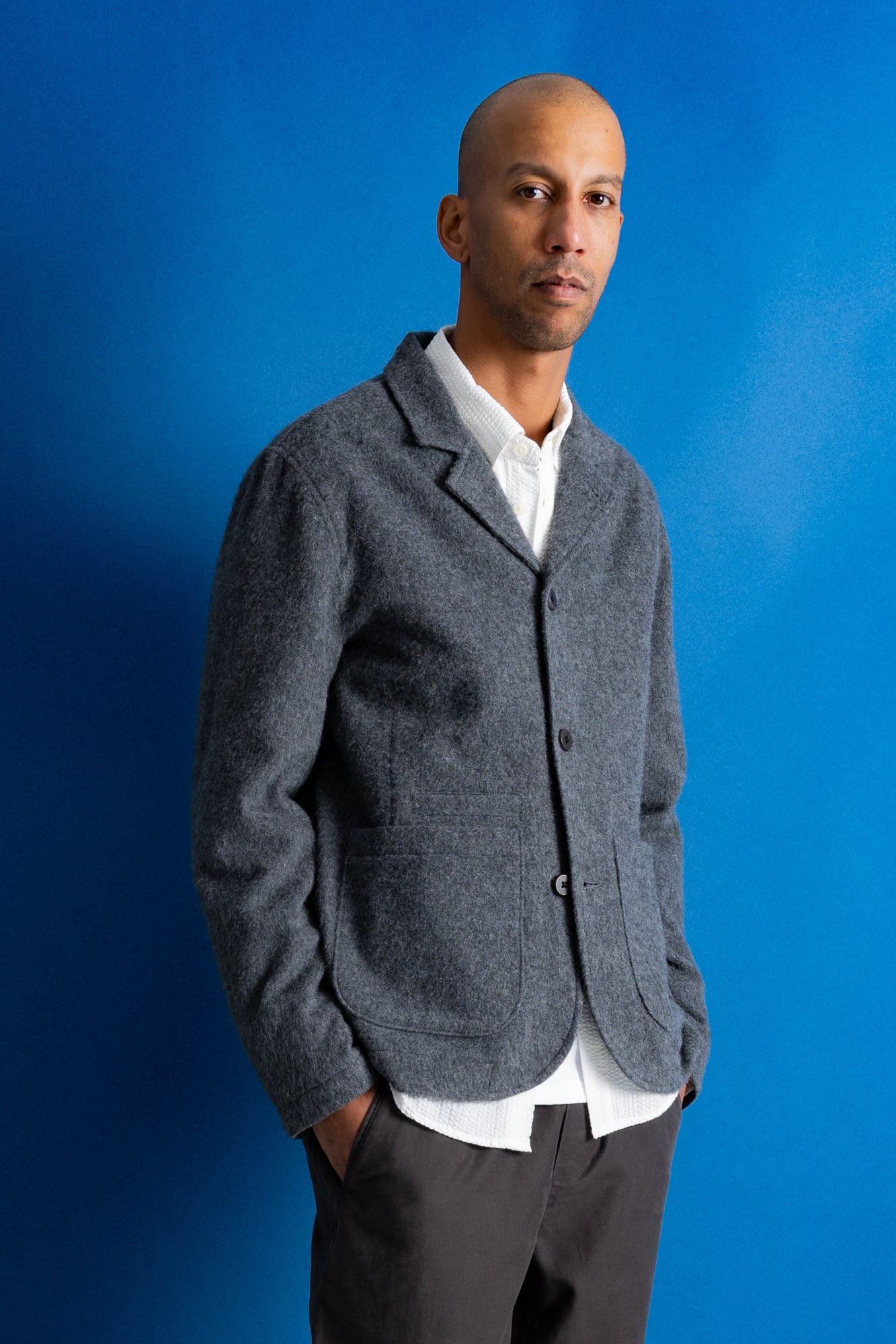 A man wearing a textured gray wool-blend blazer over a white collared shirt and dark trousers, standing against a solid blue studio background.