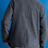 A man viewed from behind wearing a dark gray textured wool-blend jacket over a white undershirt and dark trousers, set against a solid blue studio background.