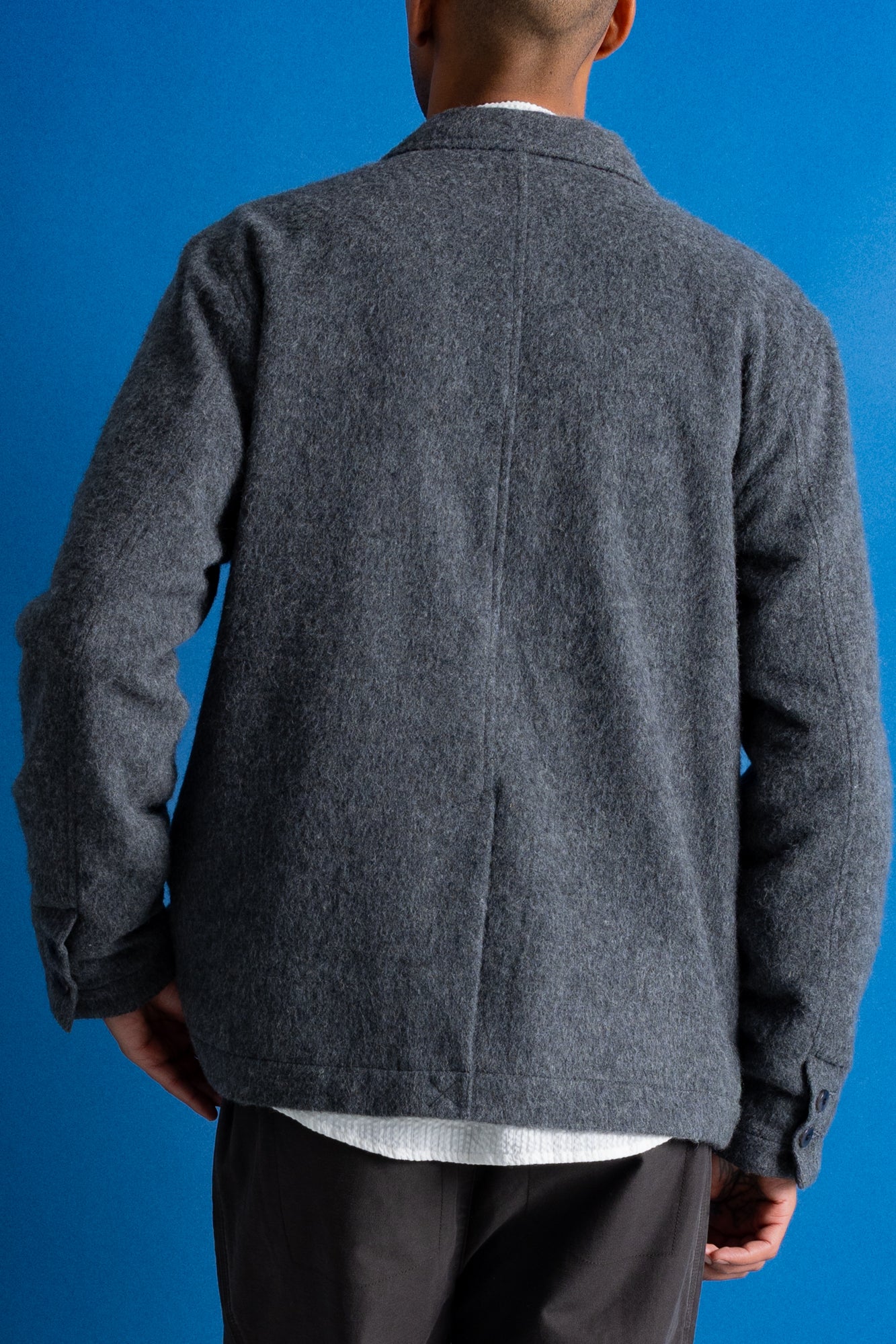 A man viewed from behind wearing a dark gray textured wool-blend jacket over a white undershirt and dark trousers, set against a solid blue studio background.