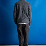 A man viewed from behind wearing a dark gray knit cardigan over a white undershirt, dark trousers, and black shoes, standing against a bright blue studio backdrop.