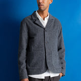 A man wearing a dark gray textured wool-blend cardigan over a white collared shirt and dark trousers, standing against a solid blue studio background.