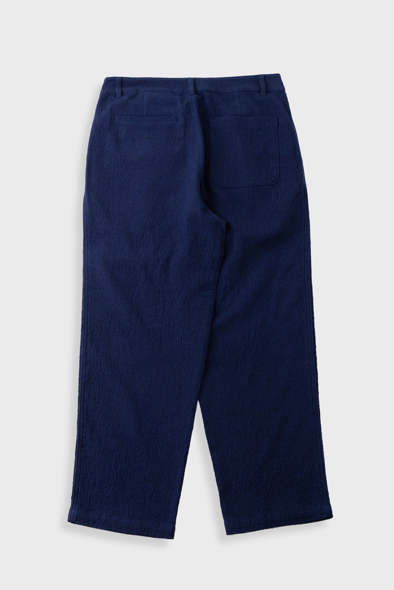 A pair of navy blue trousers shown from the back, featuring a textured crinkle fabric and a single welt pocket on the right side, set against a light gray background.