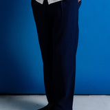 A person wearing dark navy blue relaxed-fit trousers, a white textured button-up shirt, and black leather shoes, standing against a solid blue wall.