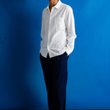 A man wearing a white long-sleeve button-down shirt and dark navy trousers, standing against a solid blue studio backdrop.