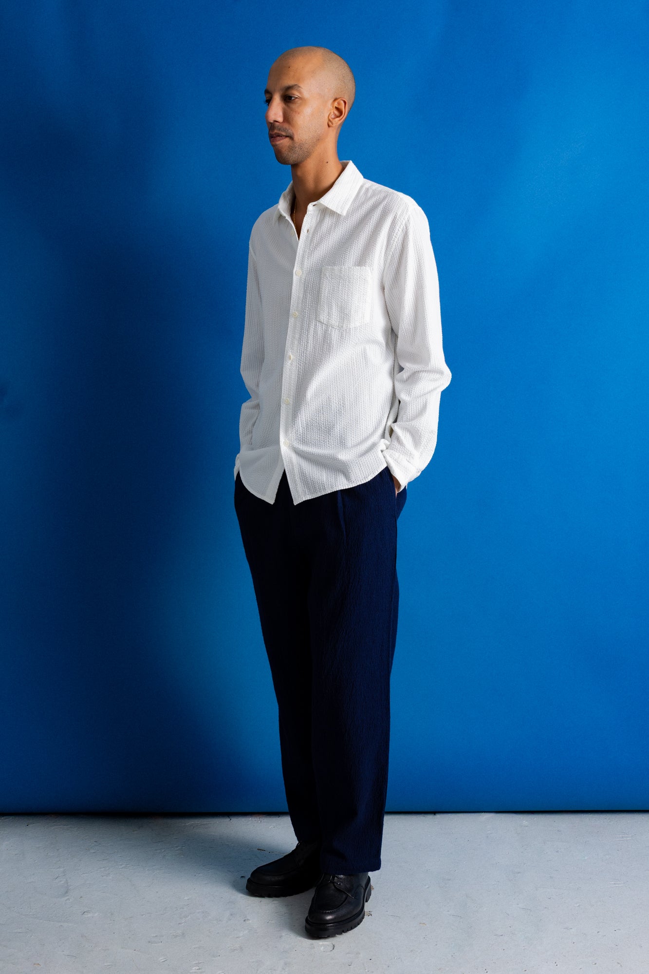 A man wearing a white long-sleeve button-down shirt and dark navy trousers, standing against a solid blue studio backdrop.