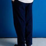A person shown from the back wearing loose dark navy trousers, a white textured shirt, and black shoes, standing against a bright blue wall on a light gray floor.