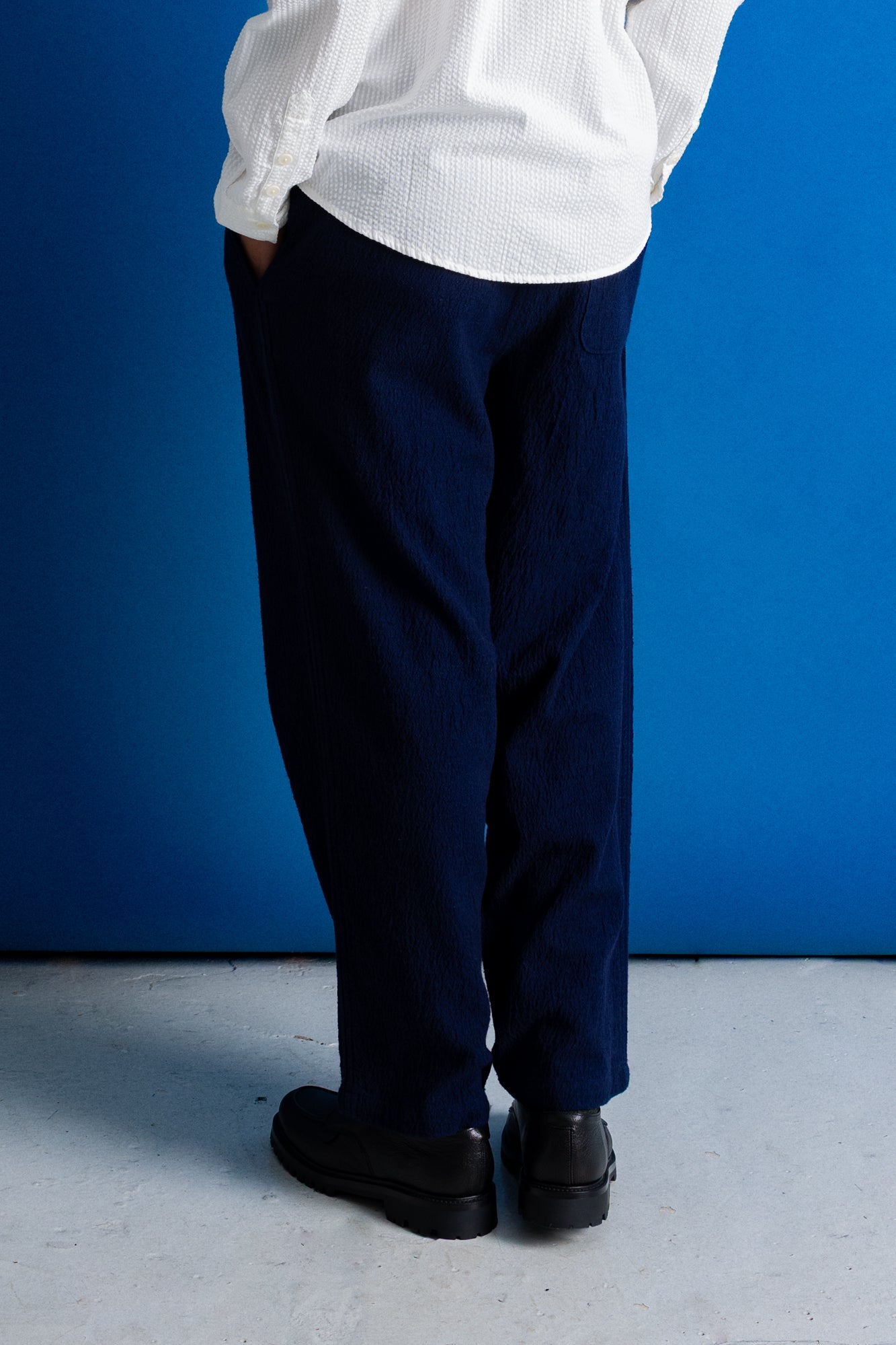 A person shown from the back wearing loose dark navy trousers, a white textured shirt, and black shoes, standing against a bright blue wall on a light gray floor.