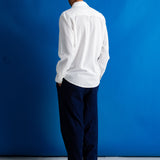 A man stands against a bright blue studio backdrop wearing a white long-sleeve button-up shirt, dark navy trousers, and black shoes.