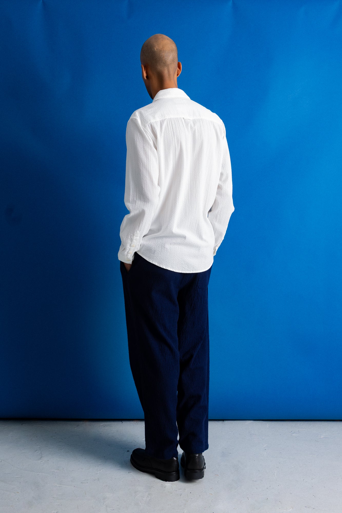A man stands against a bright blue studio backdrop wearing a white long-sleeve button-up shirt, dark navy trousers, and black shoes.