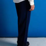 A person shown from the waist down wearing dark navy blue corduroy trousers, a white textured knit shirt, and black leather shoes, standing against a vibrant blue wall.