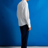 A man stands in profile wearing a white long-sleeve button-up shirt, dark navy trousers, and black shoes against a bright blue studio backdrop.