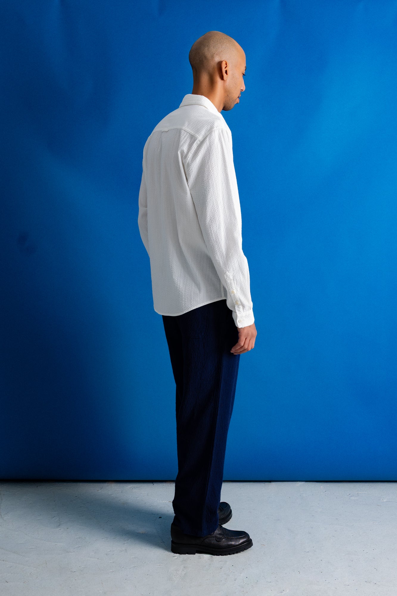 A man stands in profile wearing a white long-sleeve button-up shirt, dark navy trousers, and black shoes against a bright blue studio backdrop.