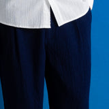 A man stands against a bright blue studio backdrop wearing a white long-sleeve button-up shirt, dark navy trousers, and black shoes.