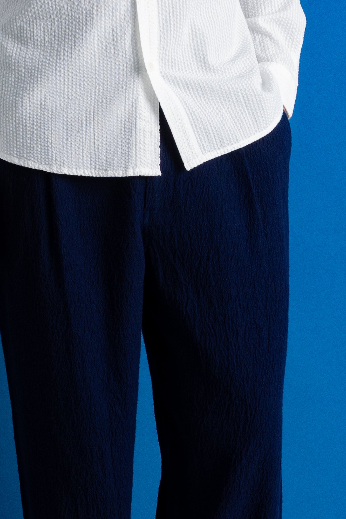 A man stands against a bright blue studio backdrop wearing a white long-sleeve button-up shirt, dark navy trousers, and black shoes.