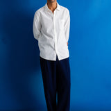 A man stands against a bright blue studio backdrop wearing a white long-sleeve button-up shirt, dark navy trousers, and black shoes.