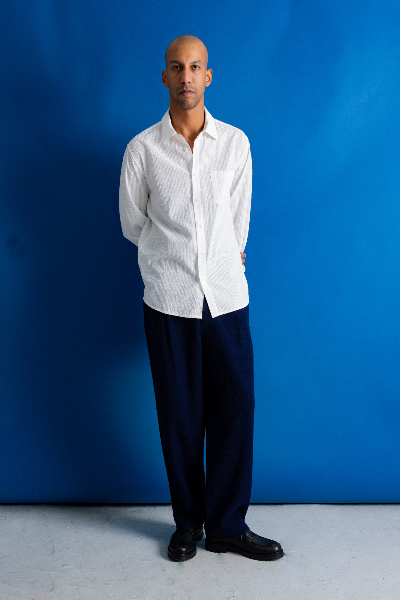 A man stands against a bright blue studio backdrop wearing a white long-sleeve button-up shirt, dark navy trousers, and black shoes.