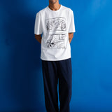 A man stands against a bright blue studio backdrop wearing a white short-sleeve T-shirt with a black comic-style graphic print on the front, dark navy pants, and black shoes.