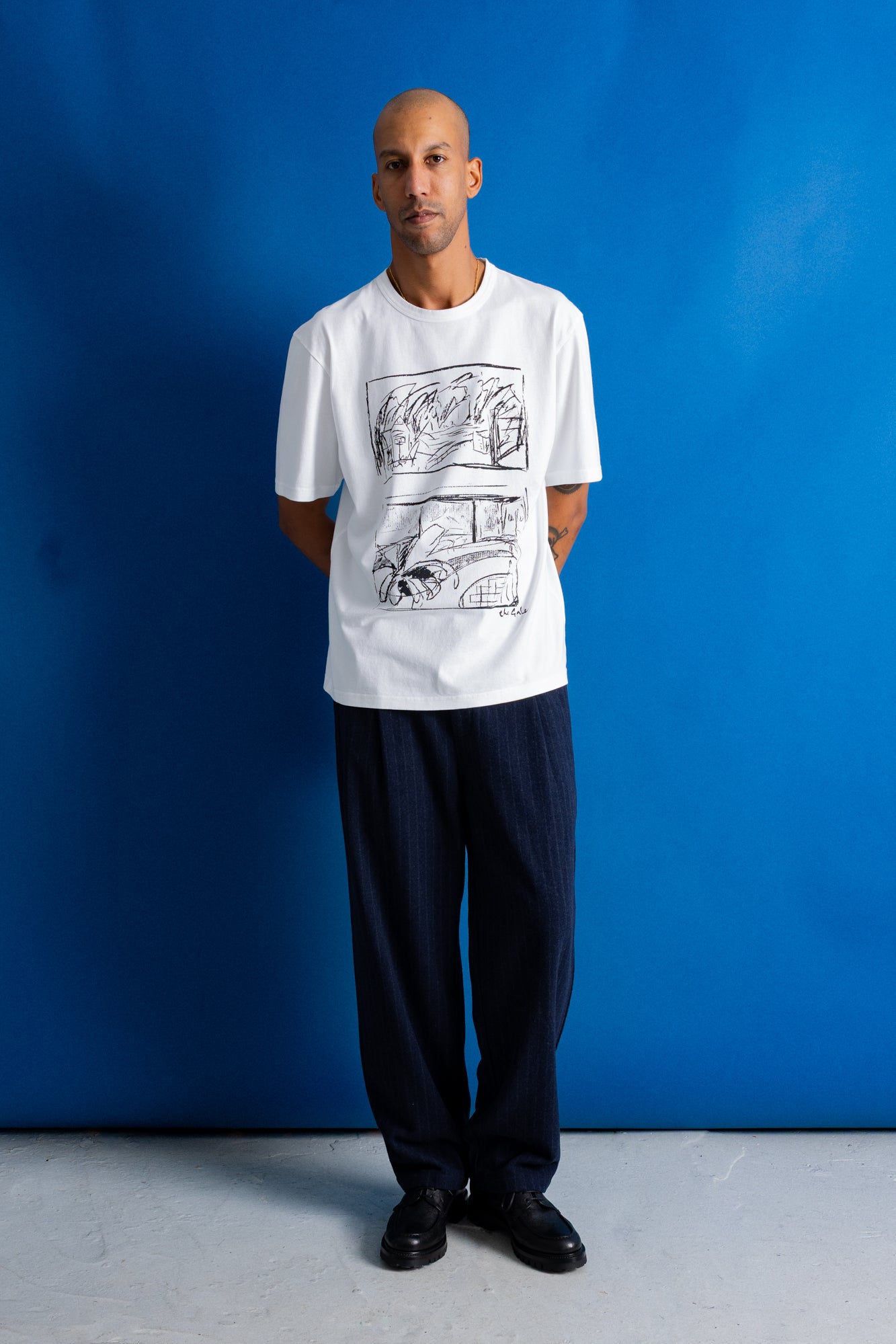 A man stands against a bright blue studio backdrop wearing a white short-sleeve T-shirt with a black comic-style graphic print on the front, dark navy pants, and black shoes.