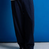 A person from the waist down wearing dark navy blue pinstriped trousers, a white graphic t-shirt, and black leather shoes, standing against a solid blue wall.