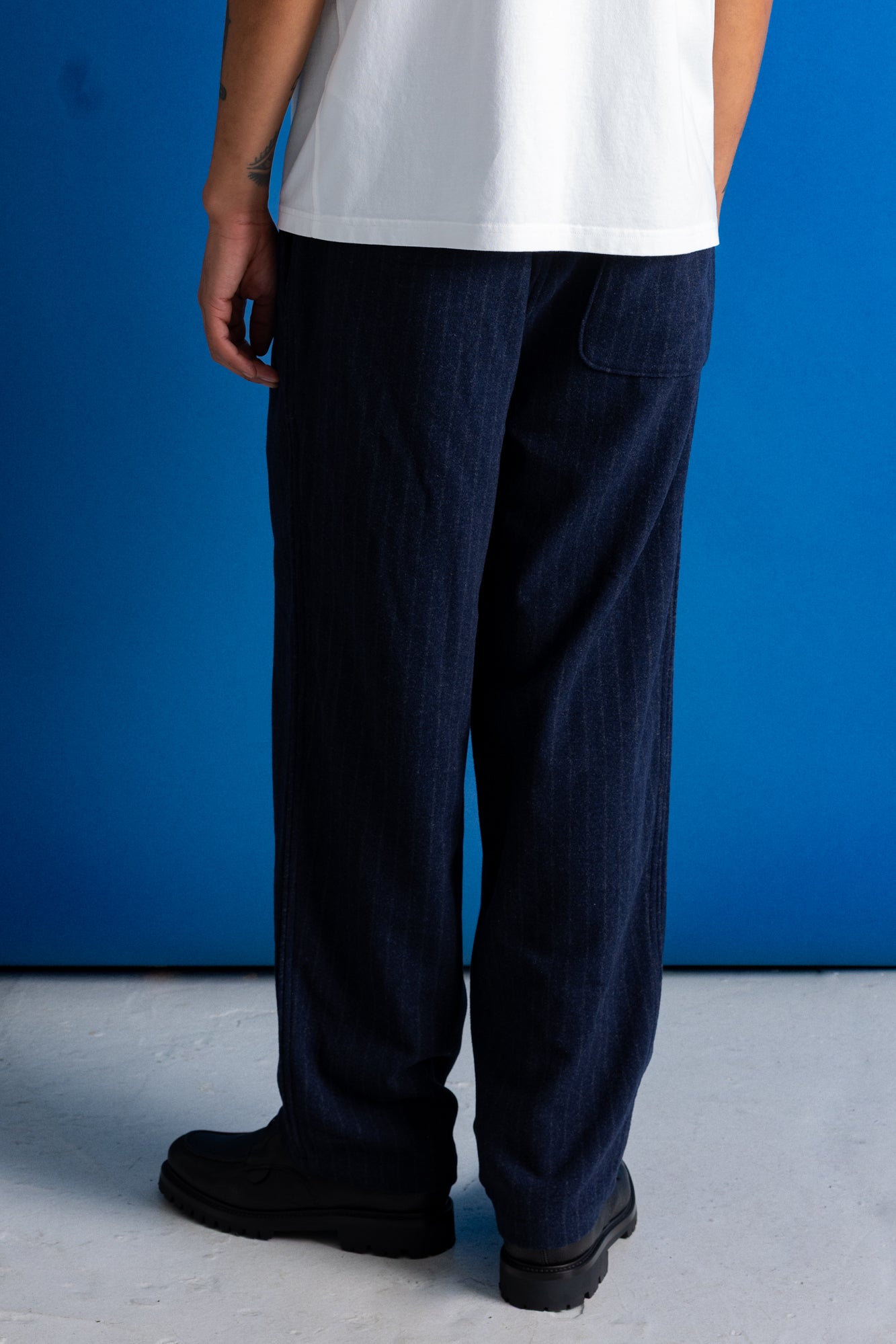 A person shown from the back wearing dark navy wide-leg denim pants, a white t-shirt, and black boots, standing against a vibrant blue studio background.
