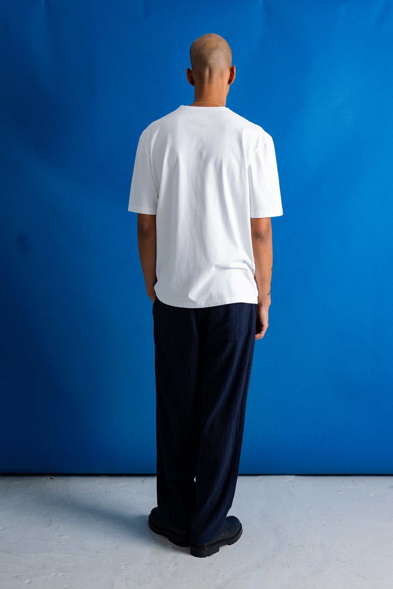 A man viewed from behind wearing a plain white short-sleeve t-shirt and dark navy wide-leg trousers, standing against a solid blue studio backdrop.