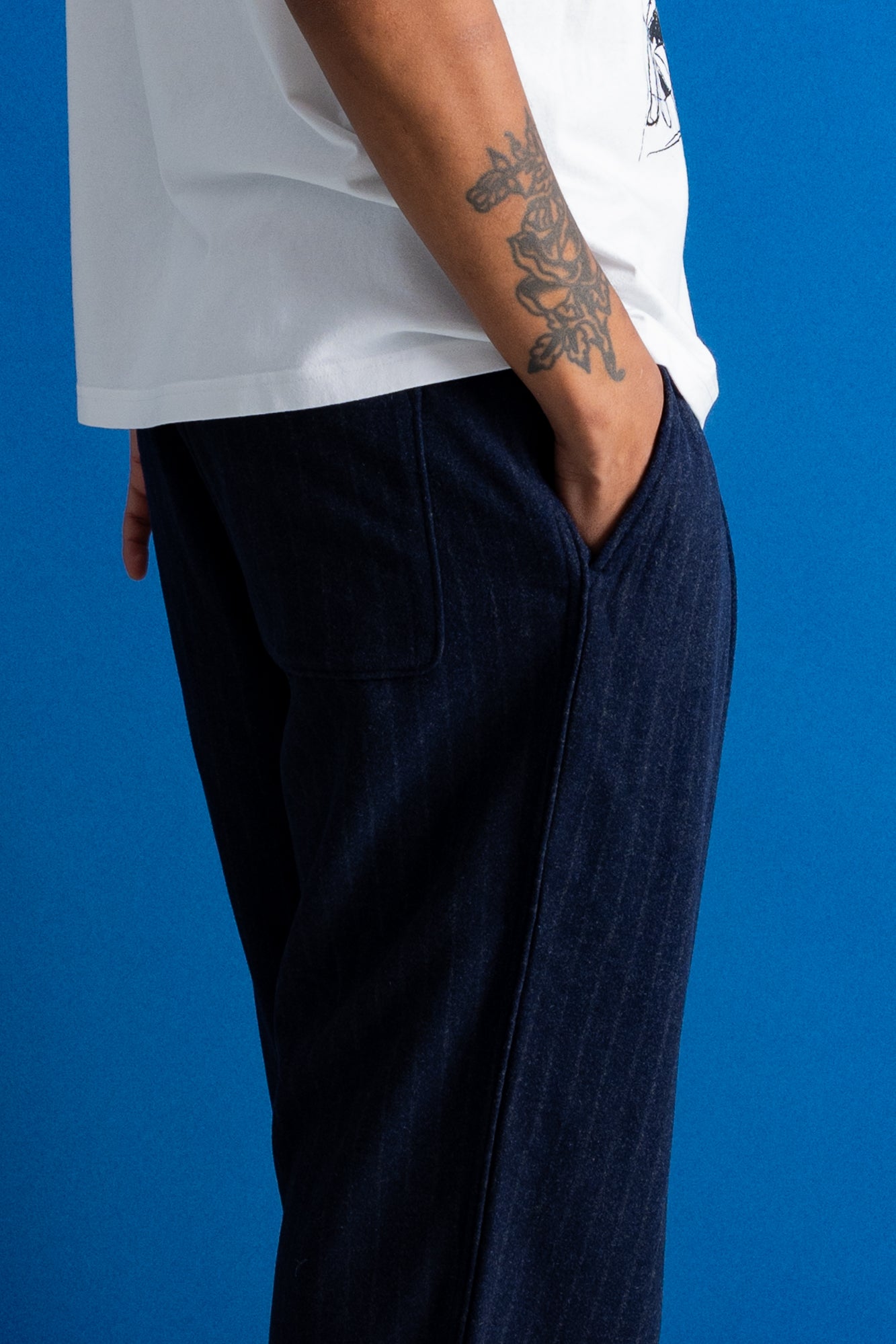 A close-up side view of a person wearing dark navy pinstriped trousers and a white t-shirt, standing against a solid blue background.