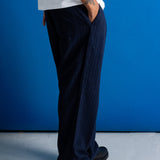 A person wearing dark navy wide-leg corduroy trousers, a white t-shirt, and black leather shoes, standing against a vibrant blue wall.