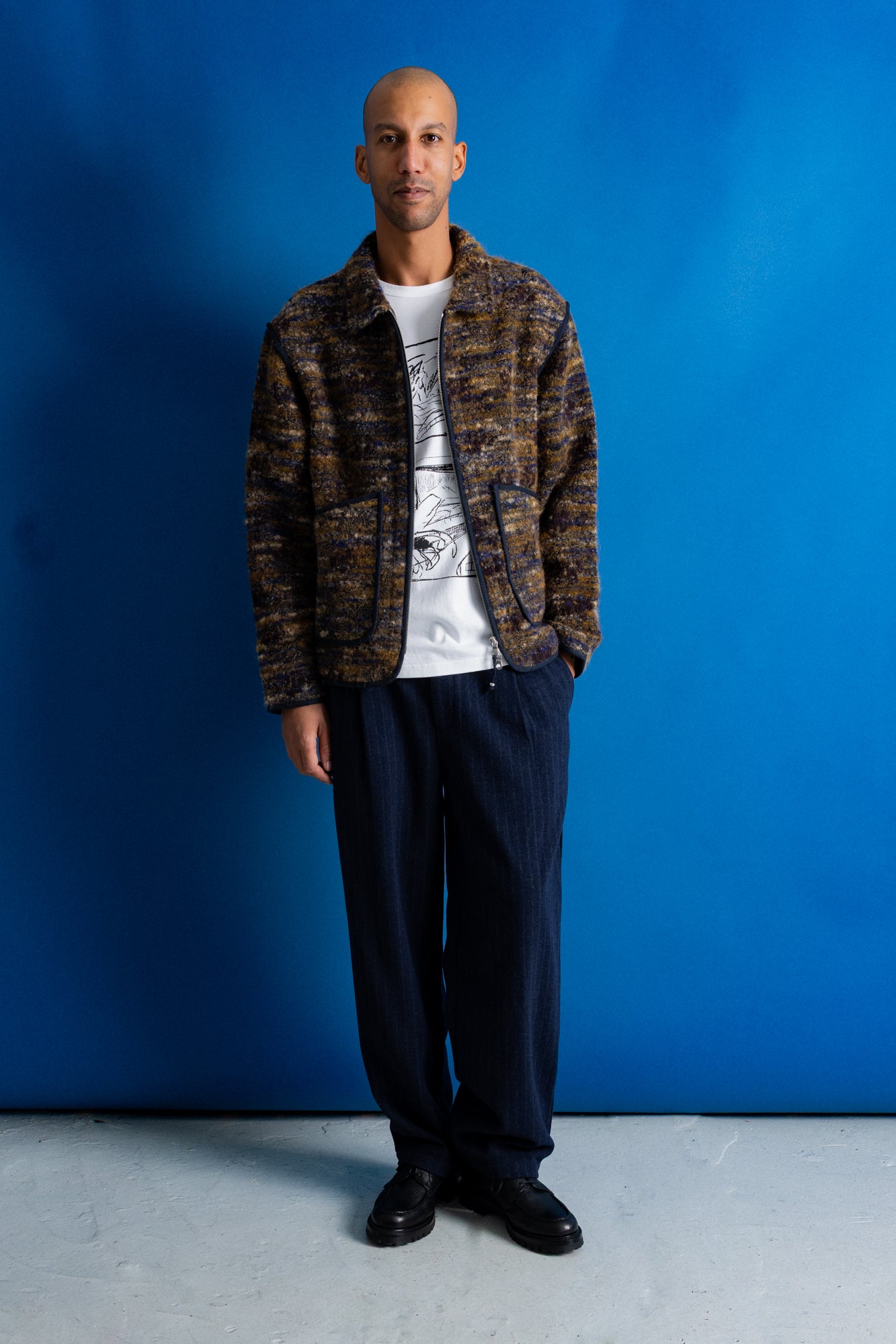 A man stands against a bright blue studio backdrop wearing a brown and black patterned zip-up jacket over a white graphic t-shirt, dark navy trousers, and black shoes.