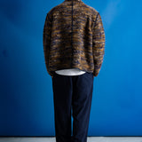 A man stands with his back to the camera wearing a textured, marled brown and gold zip-up jacket over a white t-shirt and dark navy pants, set against a solid blue studio backdrop.
