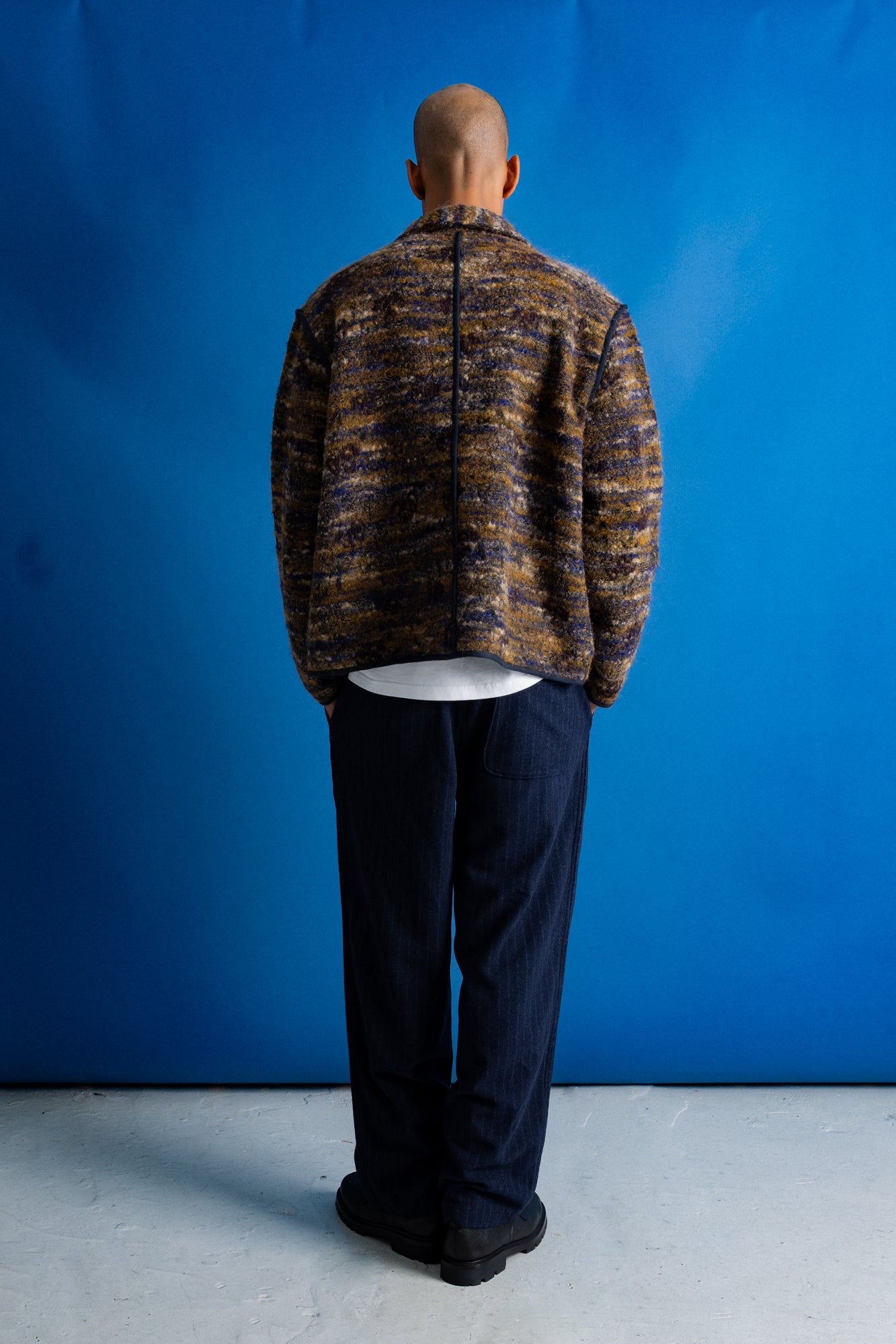 A man stands with his back to the camera wearing a textured, marled brown and gold zip-up jacket over a white t-shirt and dark navy pants, set against a solid blue studio backdrop.